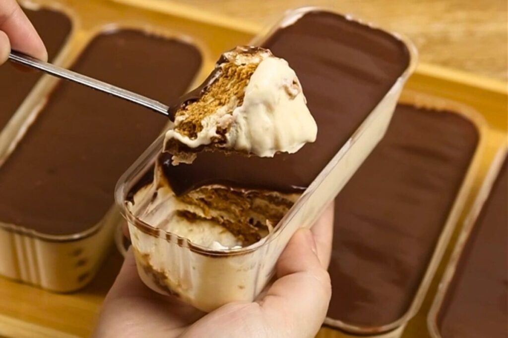 A hand scooping from a tub of Graham Boston Cream Cake, showing creamy custard layers and chocolate topping.